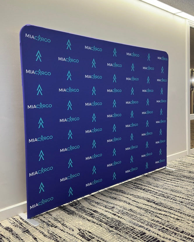 A large custom branded backdrop banner with a blue background and repeating light blue MIA/ARCO logos, set up on a carpeted floor in an indoor office or event space.