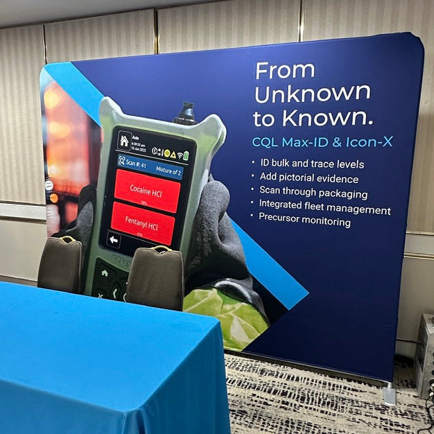 A promotional display booth for Colfax devices, featuring a large blue backdrop with product text and a handheld device mockup, alongside a blue-covered table in an indoor event space.