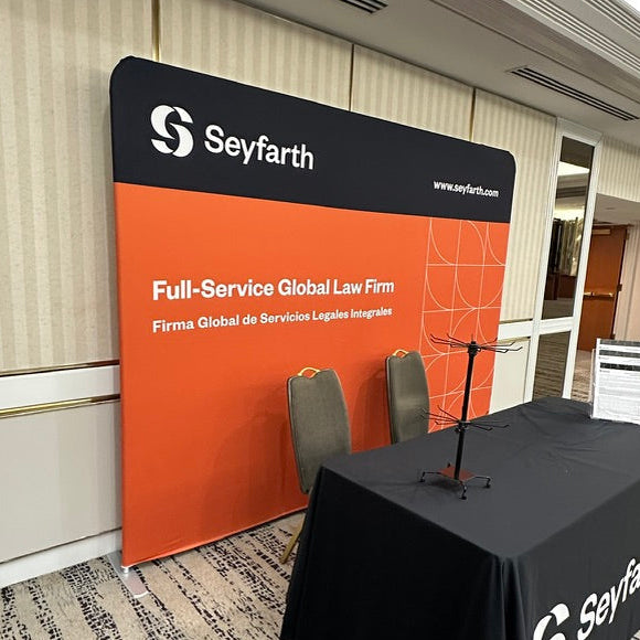 A branded Seyfarth event setup in a conference room, featuring a large orange and black backdrop panel with the Seyfarth logo and text, plus a black-covered table with matching branding.