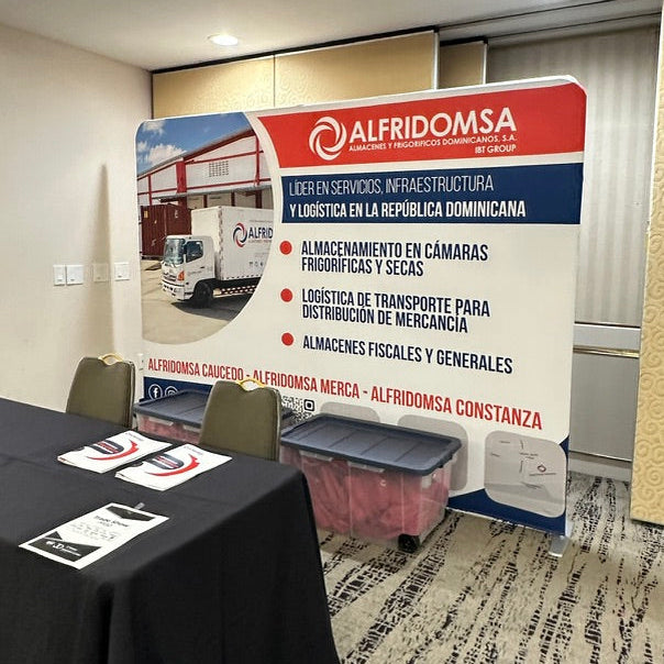 A branded event backdrop banner for 'Alfridomsa' set up in a conference room, next to a black-covered table with printed materials.
